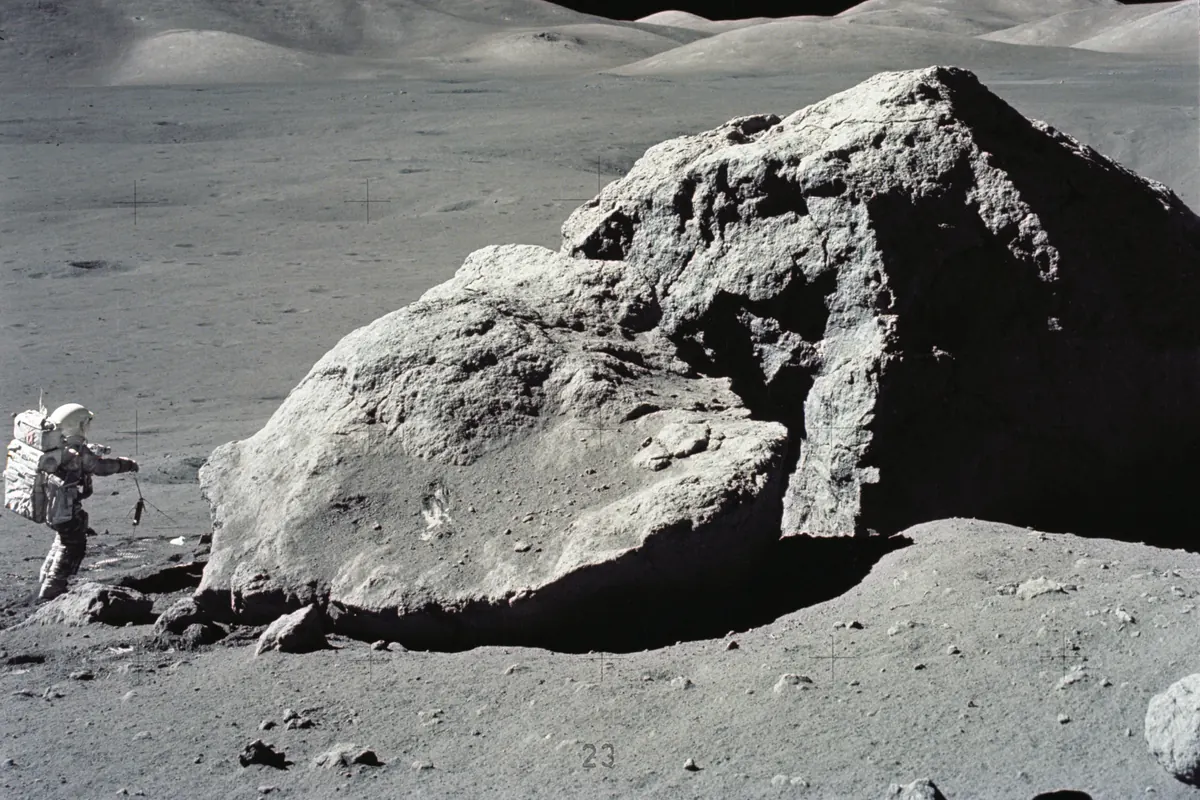 An astronaut in a white spacesuit beside a huge split boulder on the Moon