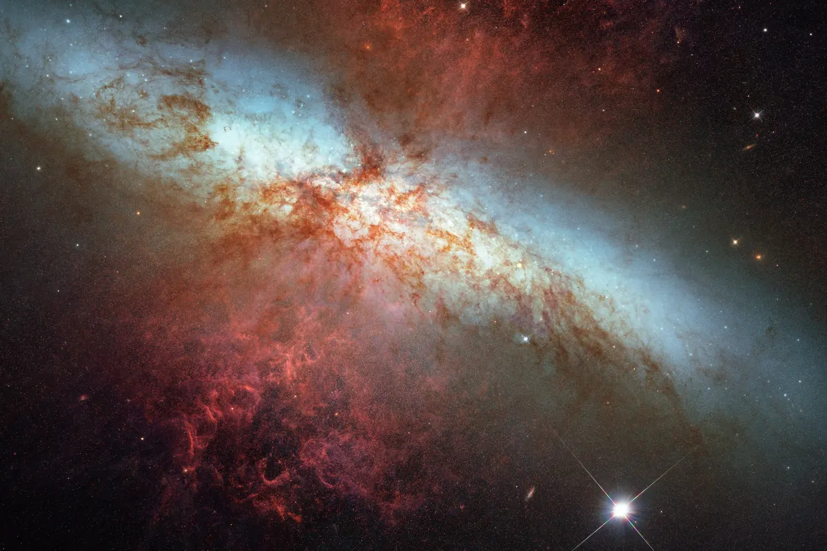 A galaxy seen edge-on with a bright blue and white disc and red gas clouds streaming from its center