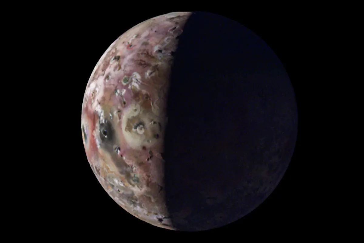 Jupiter's moon Io with a mottled surface of pink, yellow and brown, half in shadow