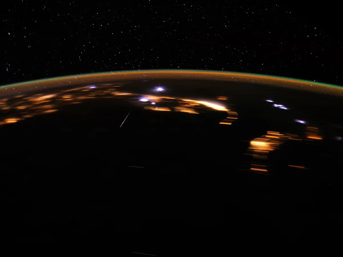 Orange city lights along Earth's dark horizon with a few meteor streaks in the atmosphere